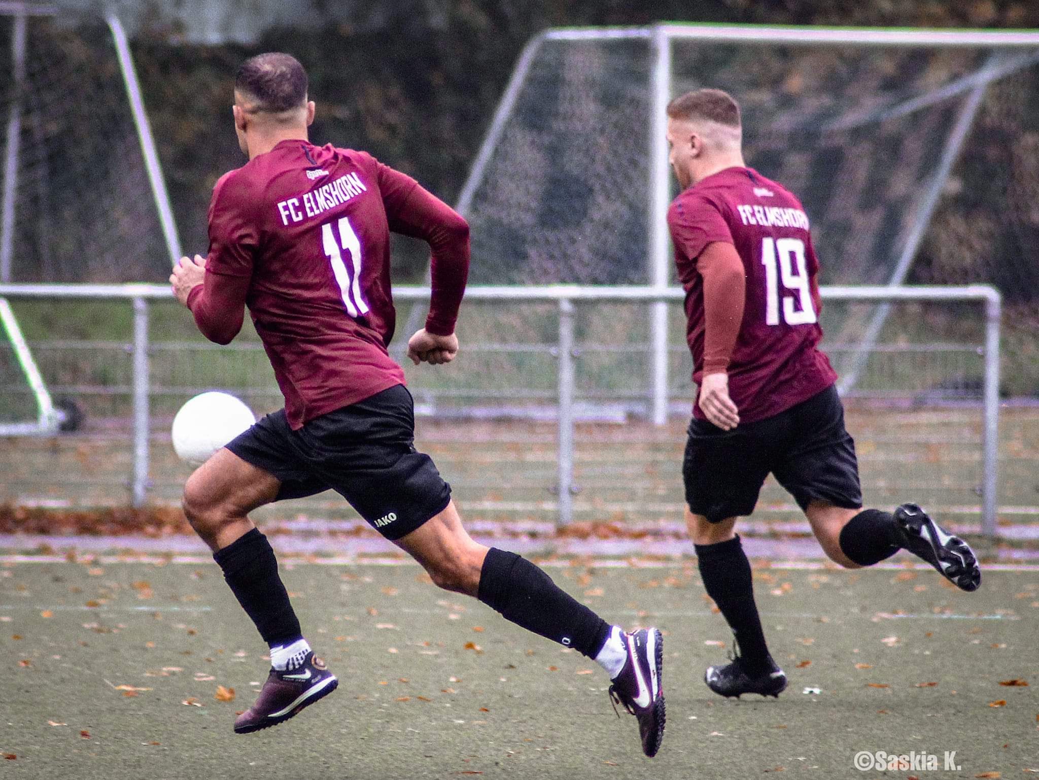 Zwei Spieler des FC Elmshorn laufen auf einem Fussballplatz mit dem Rücken zum Bild. Ein Spieler hat den Ball am Knie.